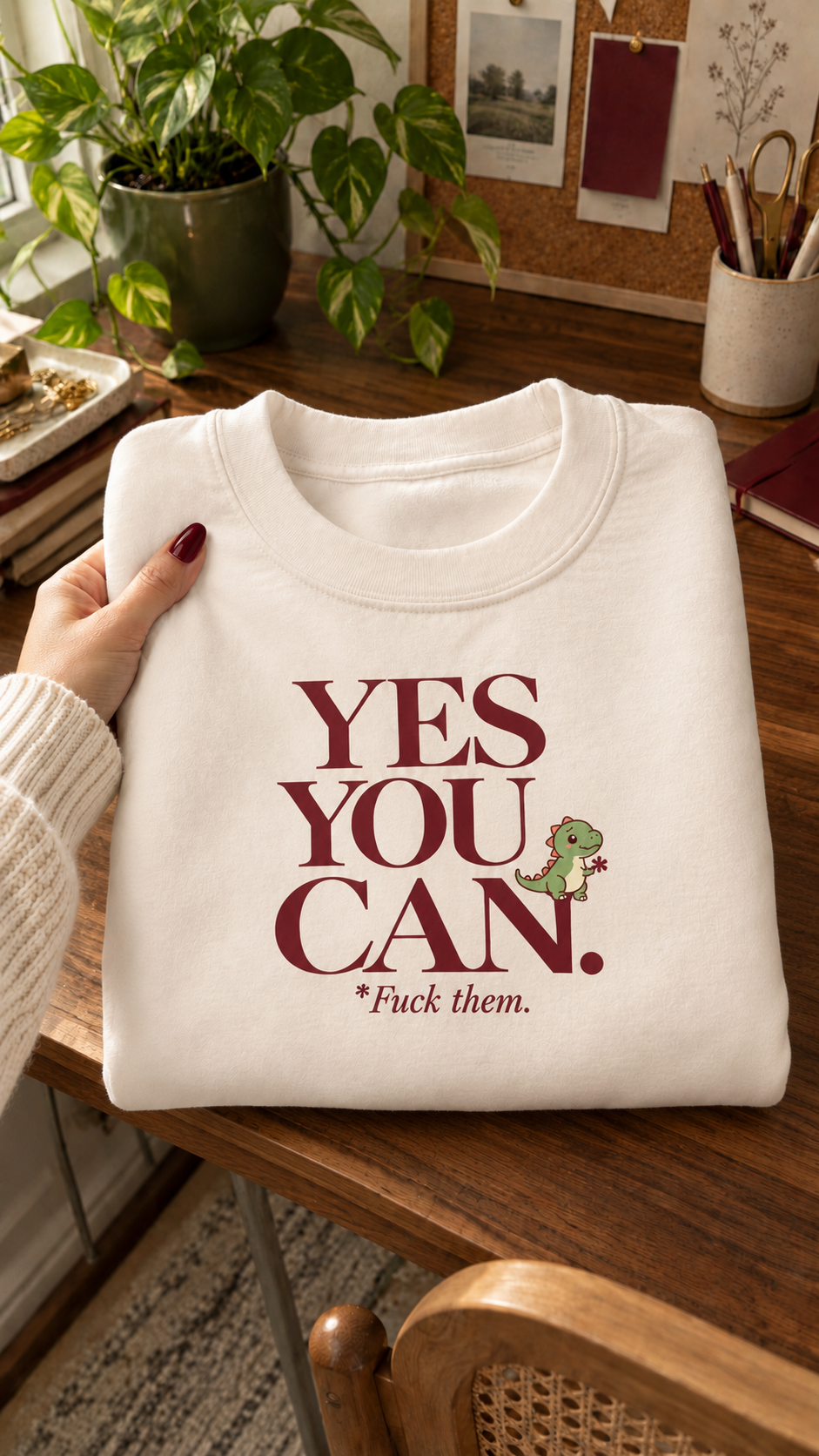 Cream Yes You Can sweatshirt with burgundy lettering and a small green dinosaur, held in a softly lit creative workspace.
